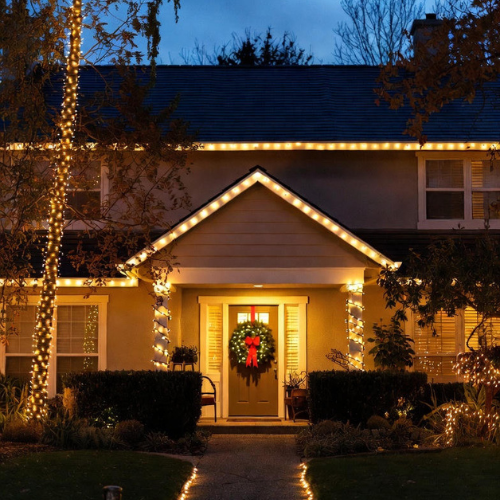 Warm white LED lights on front yard trees, roofline, and wreath creating a welcoming outdoor Christmas display.