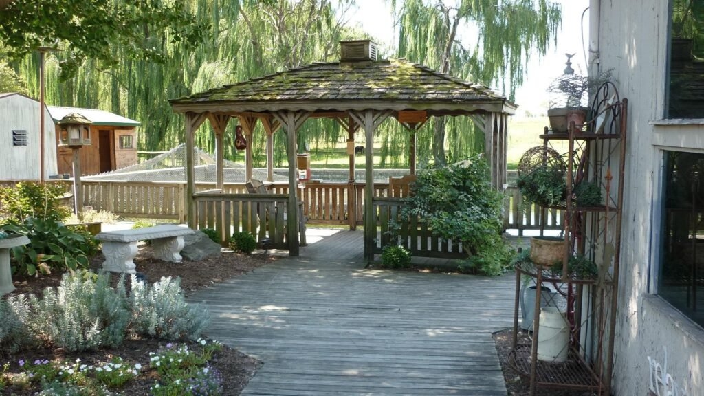 Wooden gazebo in a landscaped backyard garden surrounded by green lawn, shrubs, and flowering plants.