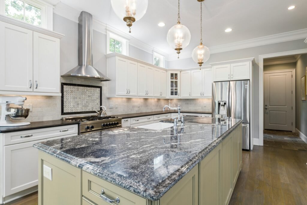 Spacious kitchen with a kitchen range exhaust hood above the cooktop, white cabinets, large island, and pendant lighting.